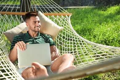 Photo of Young man with laptop resting in comfortable hammock at green garden