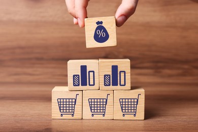 Image of Woman putting cube with percent sign on other ones with charts and shopping carts on wooden table, closeup