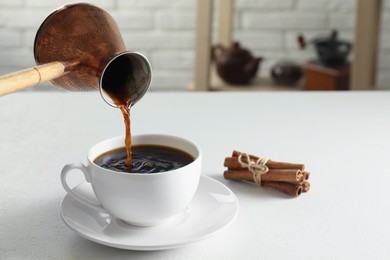 Photo of Turkish coffee. Pouring brewed beverage from cezve into cup at white table indoors