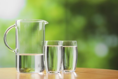 Photo of Jug and glasses with clear water on table against blurred green background, closeup. Space for text