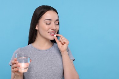 Photo of Beautiful young woman with glass of water taking pill on light blue background, space for text