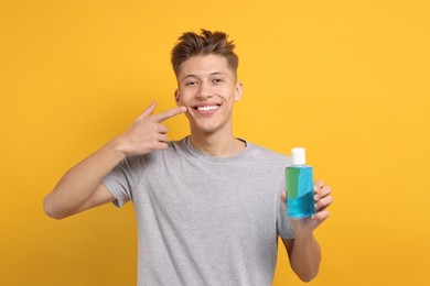 Photo of Young man with mouthwash pointing at his healthy teeth on yellow background