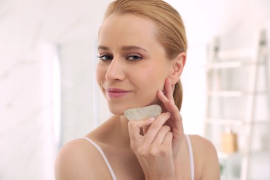 Photo of Beautiful young woman doing facial massage with gua sha tool in bathroom, closeup