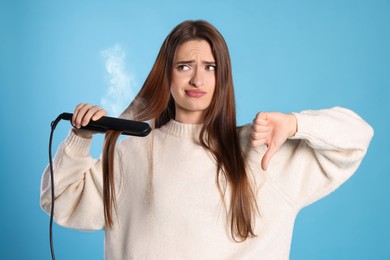 Image of Upset young woman with flattening iron showing thumb down on light blue background. Hair damage