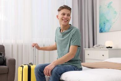 Photo of Smiling guest with suitcase relaxing on bed in stylish hotel room