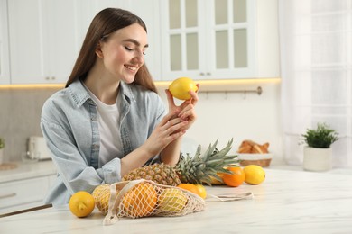 Photo of Woman with lemon and string bag of fresh fruits at light marble table in kitchen