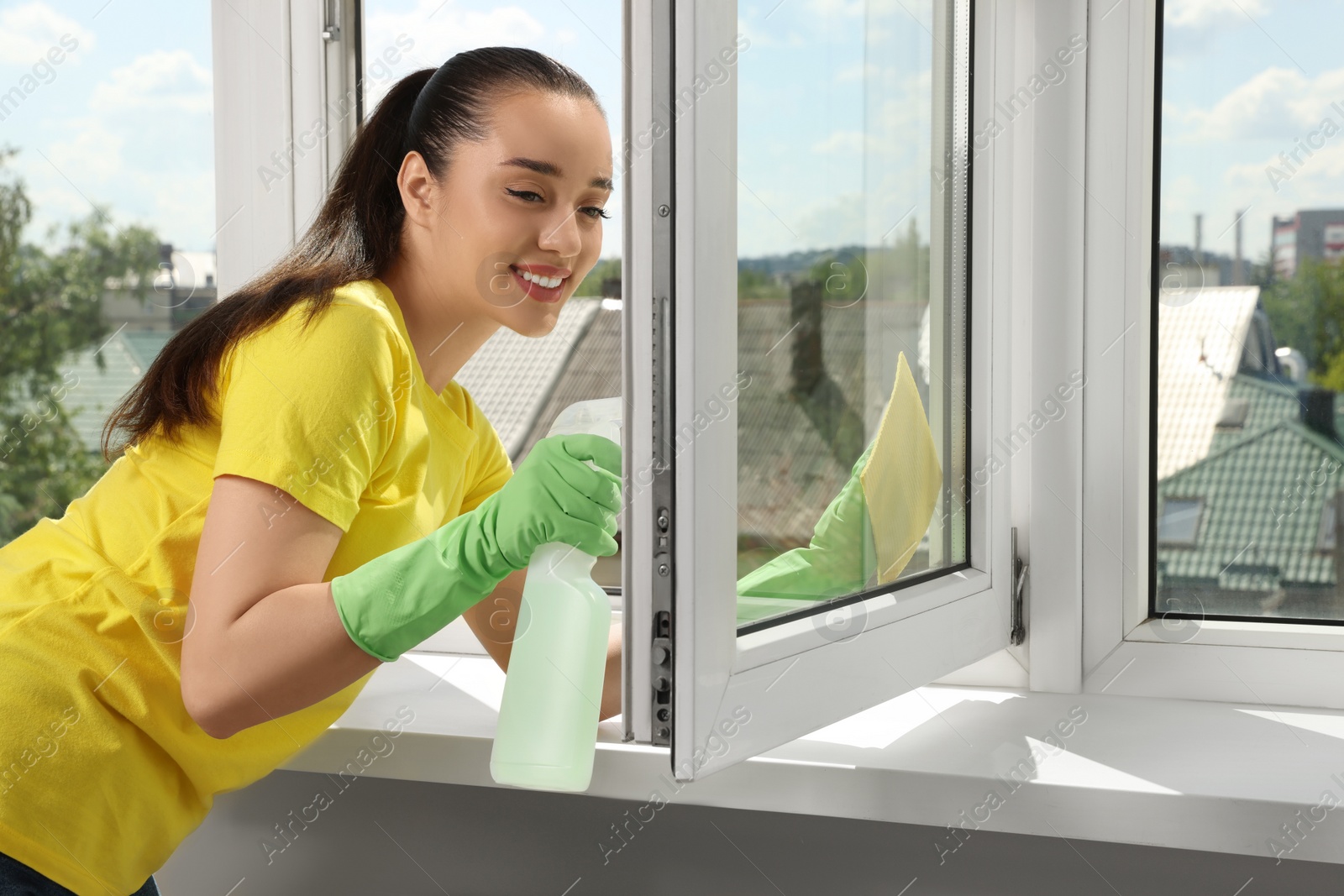 Photo of Happy young woman cleaning window glass with sponge cloth and spray indoors