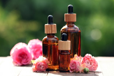 Photo of Bottles of rose essential oil and flowers on wooden table outdoors