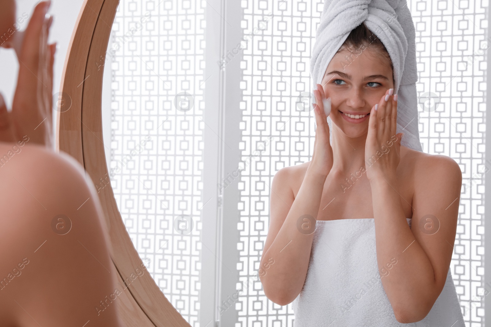 Photo of Beautiful teenage girl applying cleansing foam onto face near mirror at home. Skin care cosmetic