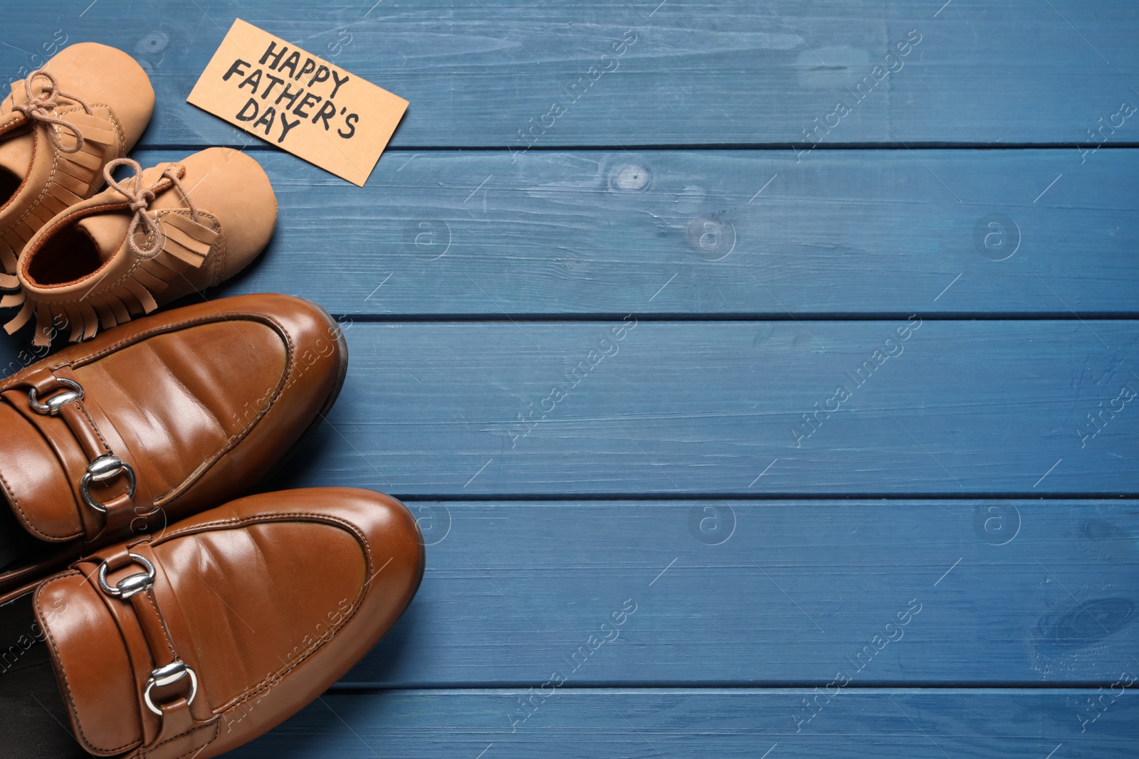 Photo of Card with phrase Happy Father's Day. Shoes for dad and child on blue wooden background, flat lay