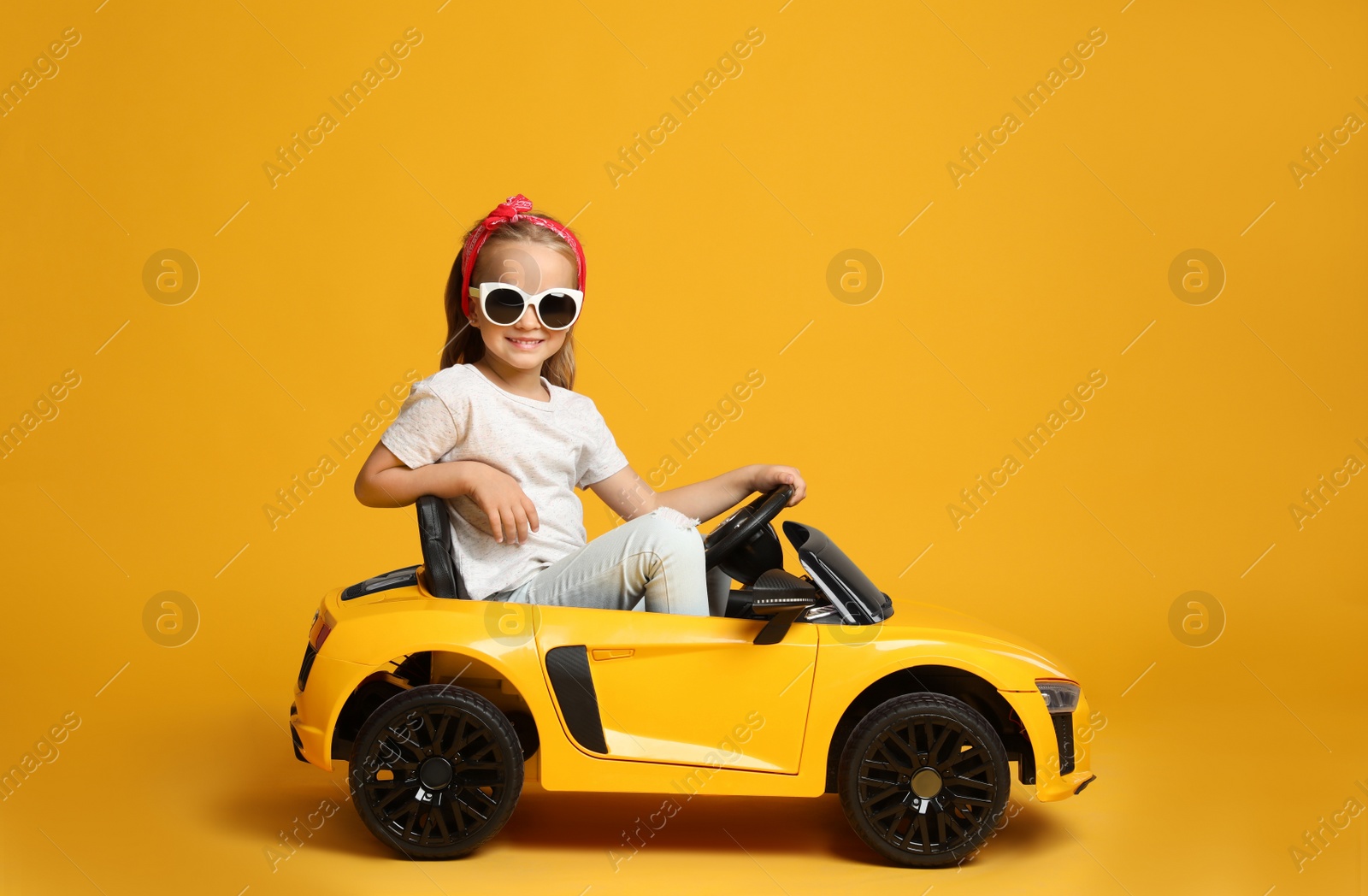 Photo of Cute little girl driving children's electric toy car on yellow background