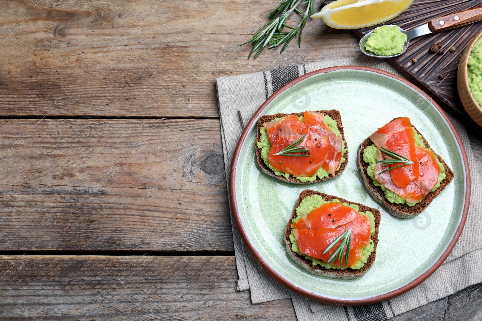 Photo of Delicious sandwiches with salmon, avocado and rosemary served on wooden table, flat lay. Space for text