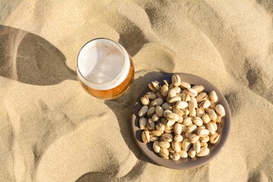 Photo of Glass of cold beer and pistachios on sandy beach, flat lay