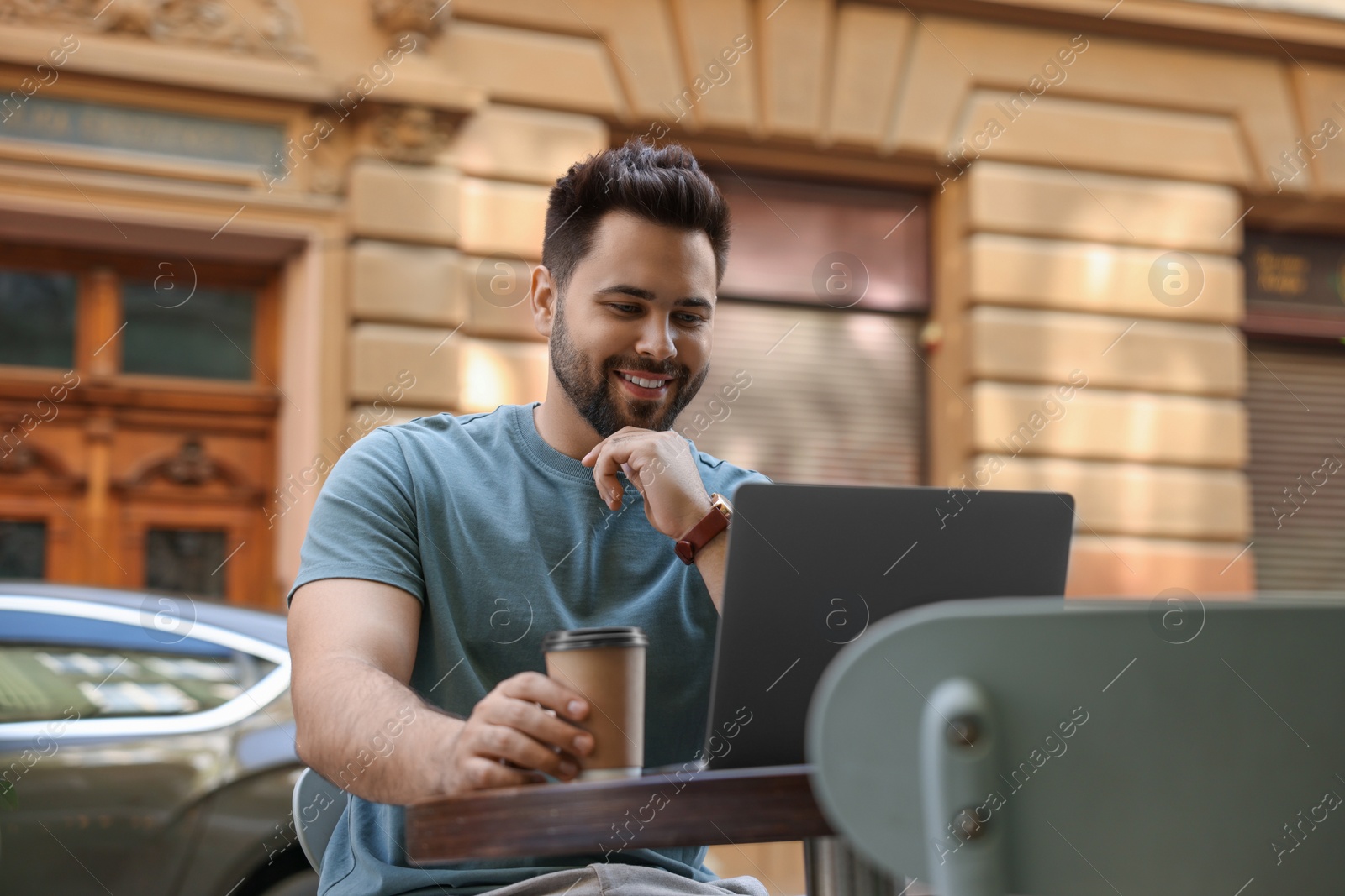 Photo of Handsome young man with cup of coffee working on laptop at table in outdoor cafe