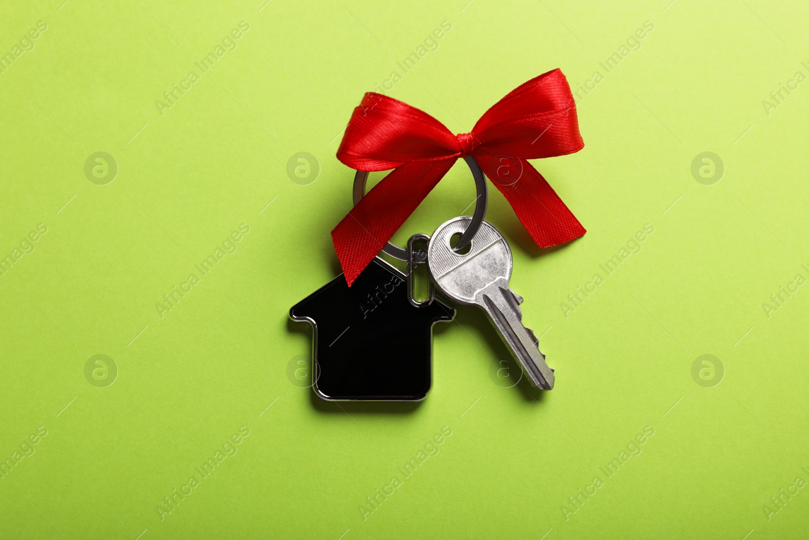 Photo of Key with trinket in shape of house and red bow on light green background, top view. Housewarming party