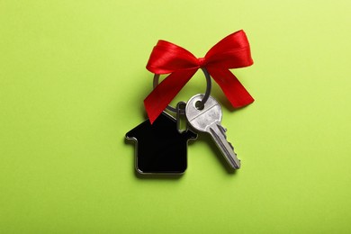 Photo of Key with trinket in shape of house and red bow on light green background, top view. Housewarming party