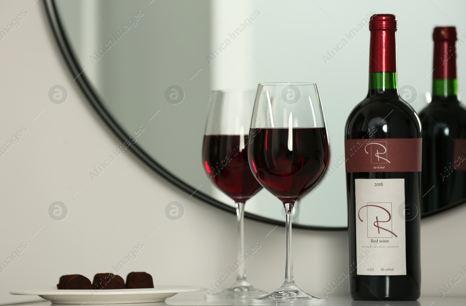 Photo of Bottle and glasses of red wine with chocolate candies on table near mirror