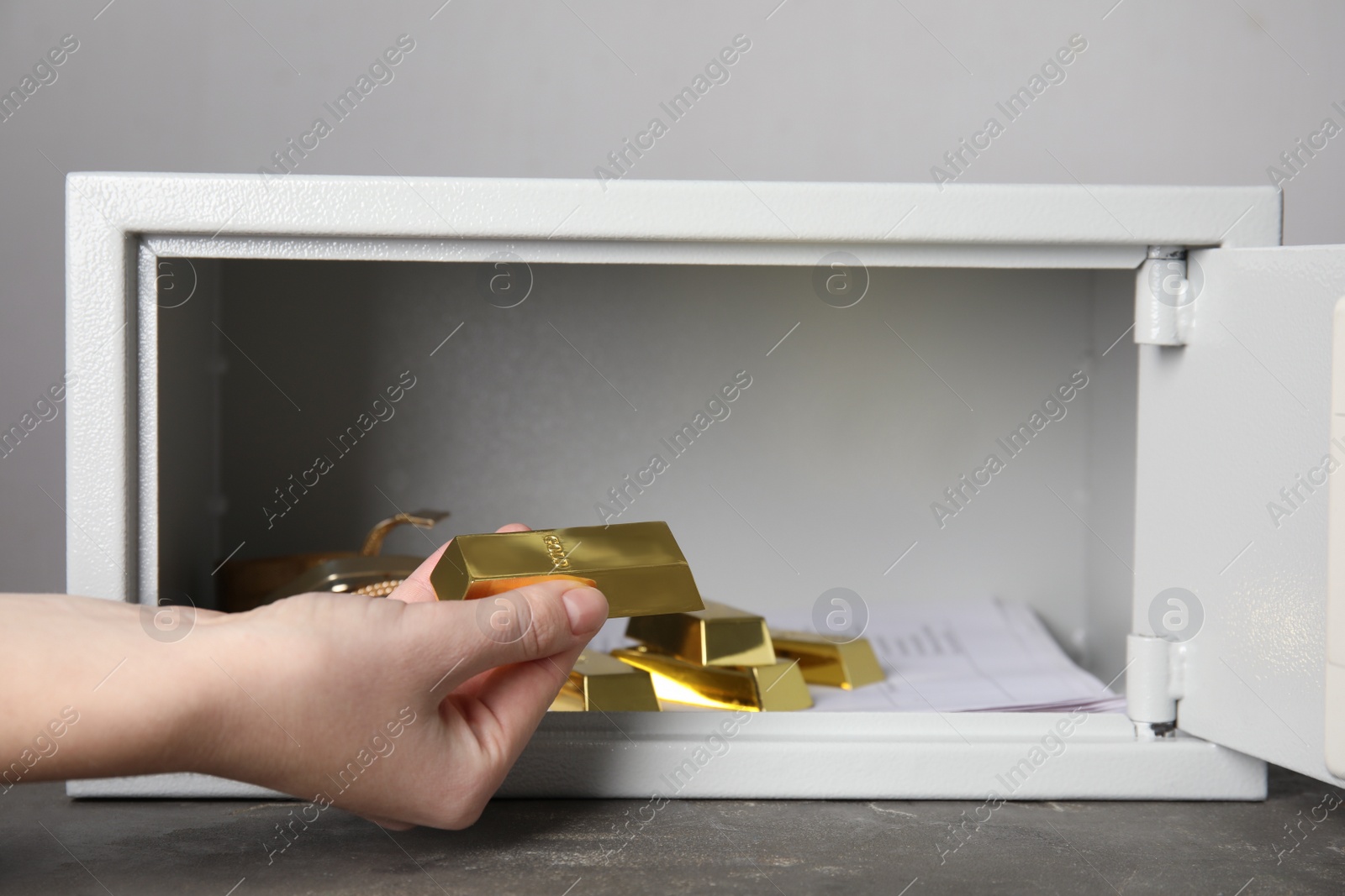 Photo of Woman taking gold bar out of steel safe on grey table against light background, closeup