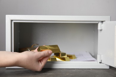 Photo of Woman taking gold bar out of steel safe on grey table against light background, closeup