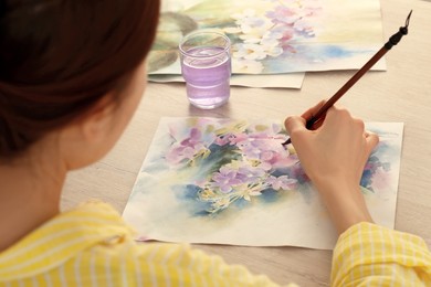 Woman painting flowers with watercolor at white wooden table, closeup. Creative artwork