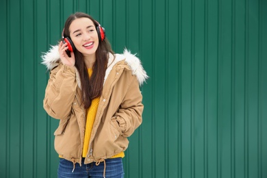 Beautiful young woman listening to music with headphones against color wall. Space for text