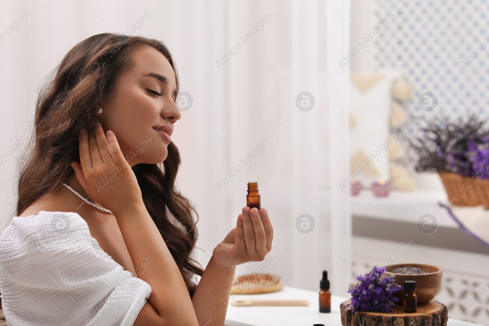 Photo of Beautiful young woman applying essential oil onto neck indoors