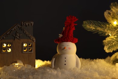 Photo of Cute decorative snowman, hut and Christmas tree on artificial snow against dark background