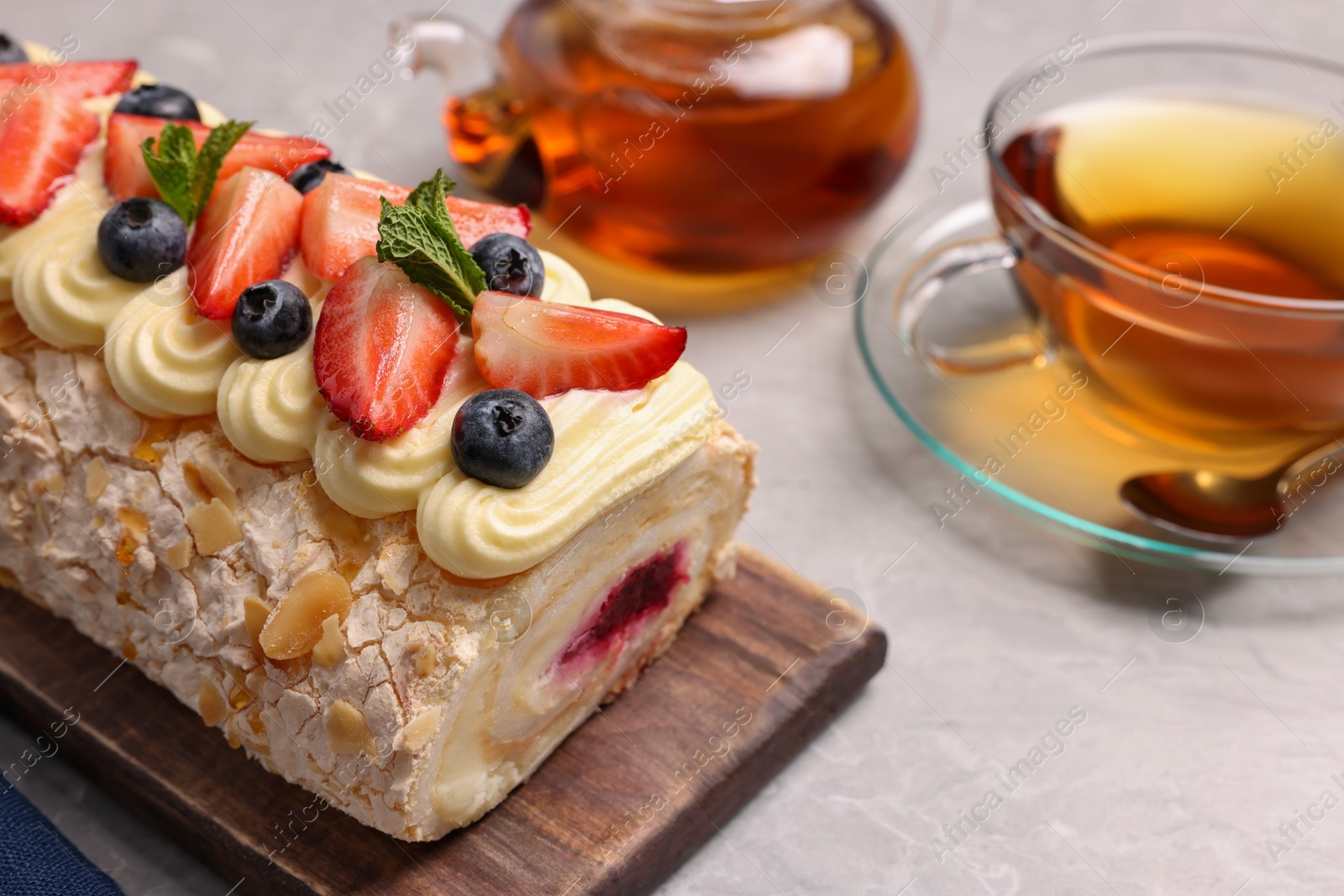 Photo of Tasty meringue roll with jam, cream, strawberry, blueberry, mint and aromatic tea on light grey table, closeup