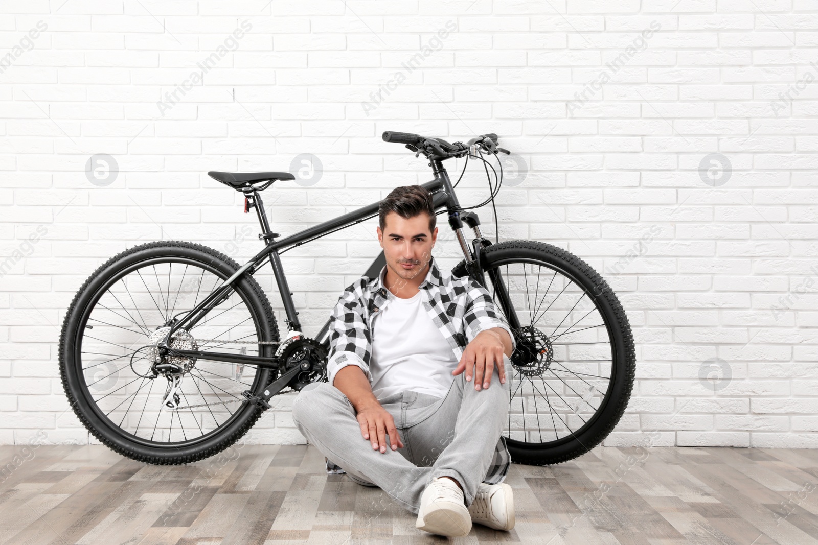 Photo of Handsome young man with modern bicycle near white brick wall indoors