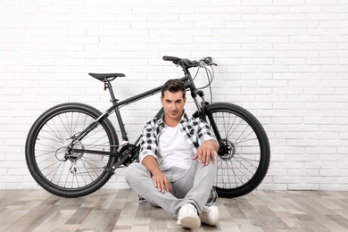 Handsome young man with modern bicycle near white brick wall indoors