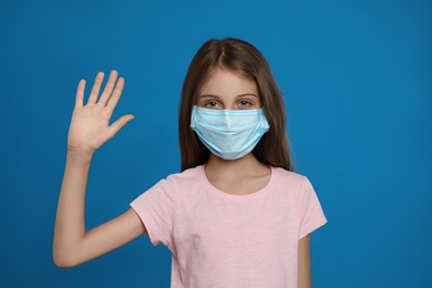 Photo of Little girl in protective mask showing hello gesture on blue background. Keeping social distance during coronavirus pandemic
