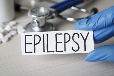 Doctor holding card with word Epilepsy at white wooden table, closeup