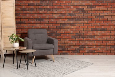 Cosy armchair and potted plant on coffee table near brick wall in room, space for text. Interior design