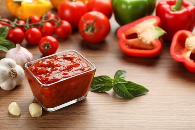 Photo of Delicious adjika sauce in glass bowl and ingredients on wooden table