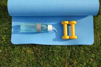 Photo of Dumbbells, bottle of water and fitness mat on green grass, flat lay. Morning exercise