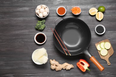 Empty iron wok and chopsticks surrounded by ingredients on dark grey wooden table, flat lay. Space for text