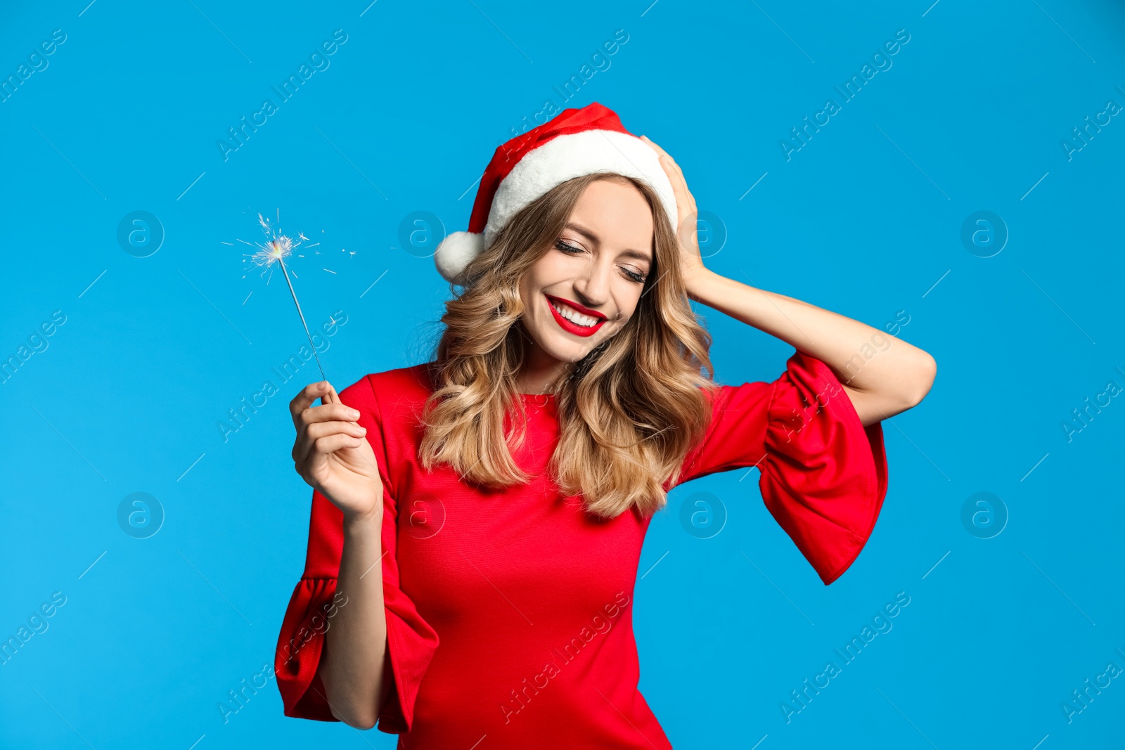 Photo of Happy young woman wearing Santa hat with sparkler on light blue background. Christmas celebration