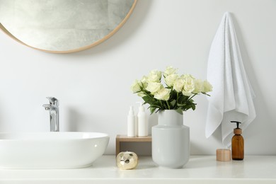 Vase with beautiful white roses and toiletries near sink in bathroom