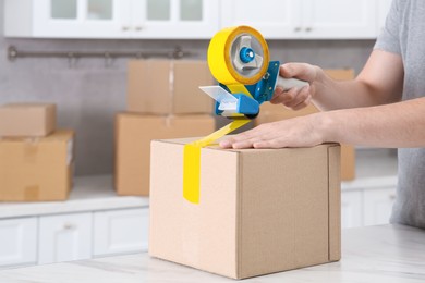 Photo of Man taping box with adhesive tape dispenser in kitchen, closeup