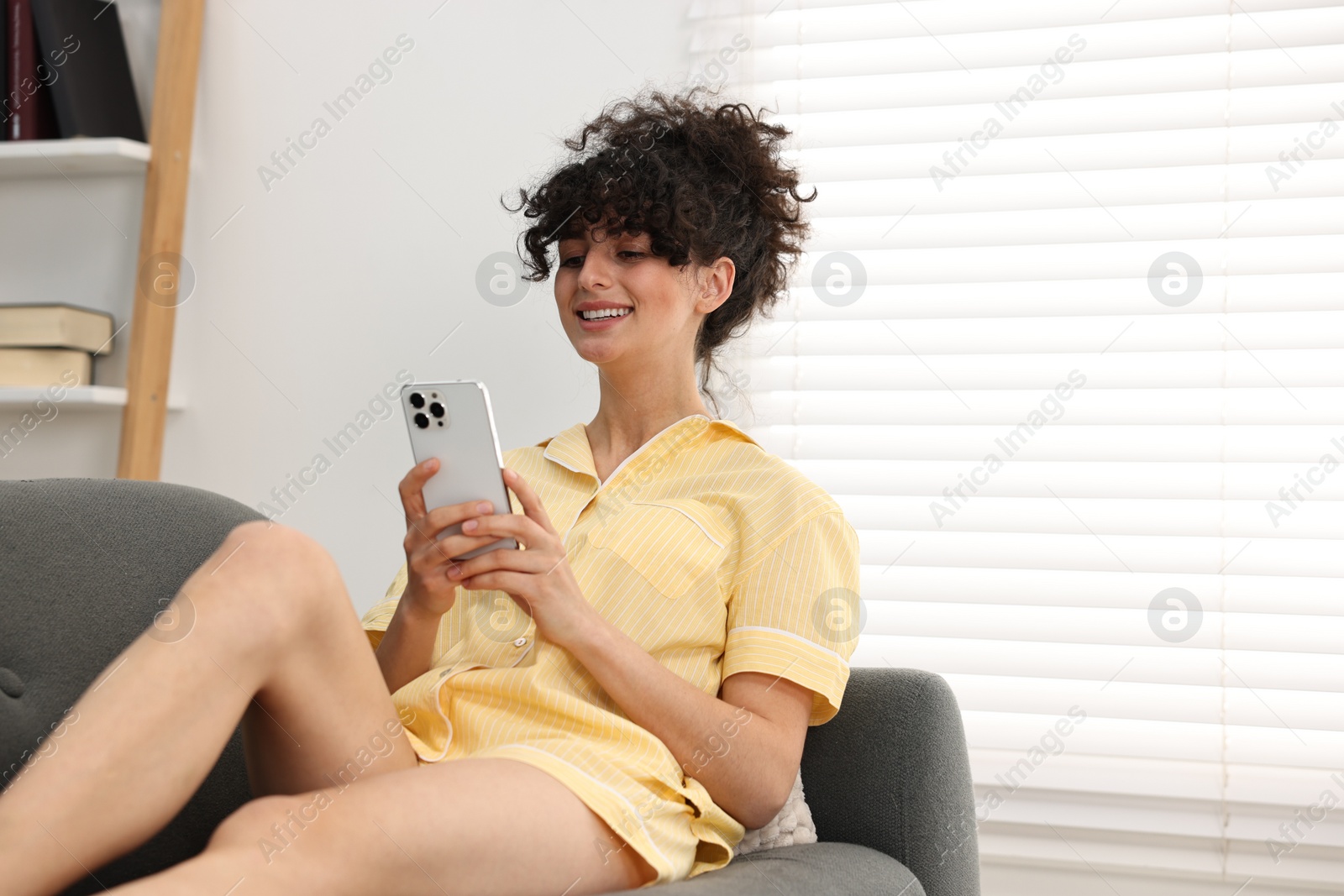 Photo of Beautiful young woman in stylish pyjama using smartphone on sofa at home