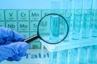 Photo of Scientist with magnifying glass examining test tube against periodic table of elements in laboratory, closeup. Color tone effect