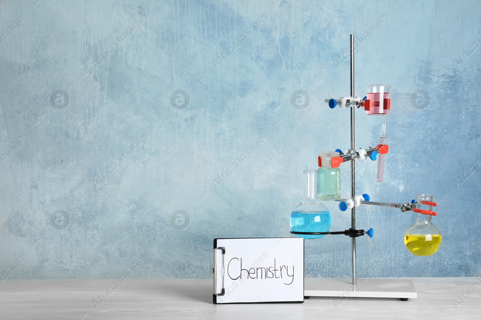 Photo of Laboratory glassware and clipboard with word CHEMISTRY on table against color background, space for text