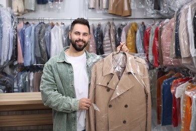 Photo of Dry-cleaning service. Happy man holding hanger with coat in plastic bag indoors