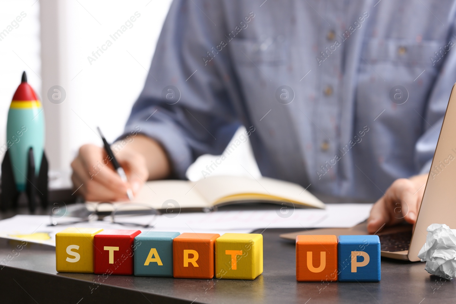 Photo of Woman working at messy table, focus on colorful cubes with words Start up