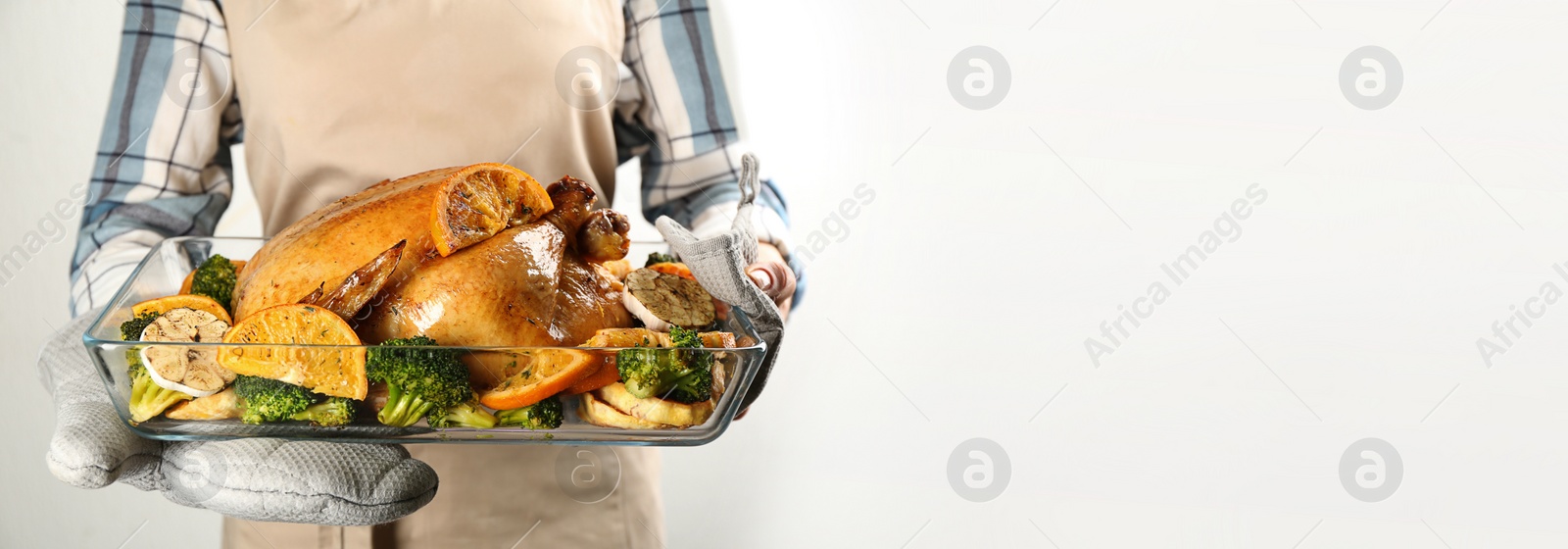 Photo of Woman holding pan with chicken, oranges and vegetables on light background, closeup