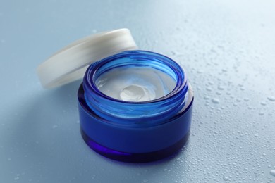 Moisturizing cream in open jar on light blue background with water drops, closeup