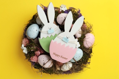 Photo of Wooden bunnies with protective masks, painted eggs and Easter wreath on yellow background, top view. Holiday during COVID-19 quarantine