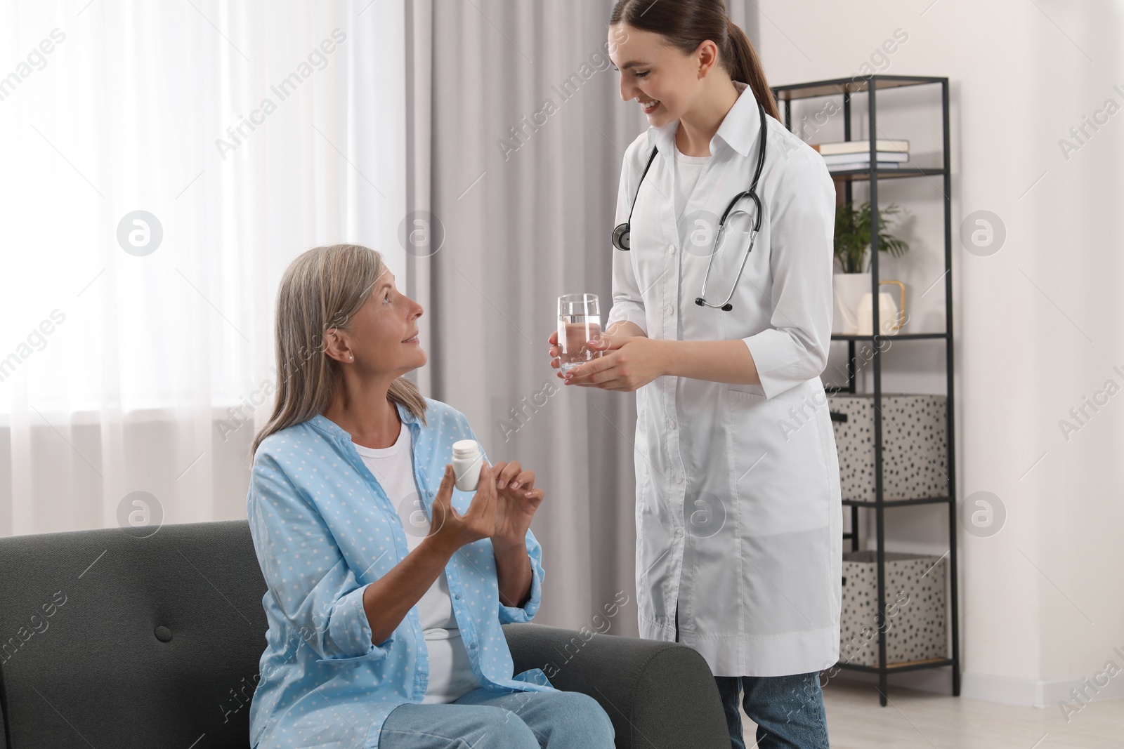 Photo of Young healthcare worker giving glass of water to senior woman with pills indoors