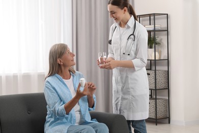 Photo of Young healthcare worker giving glass of water to senior woman with pills indoors