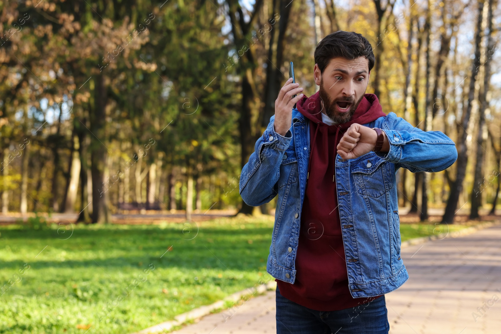 Photo of Emotional man talking by smartphone and checking time on watch in park, space for text. Being late concept